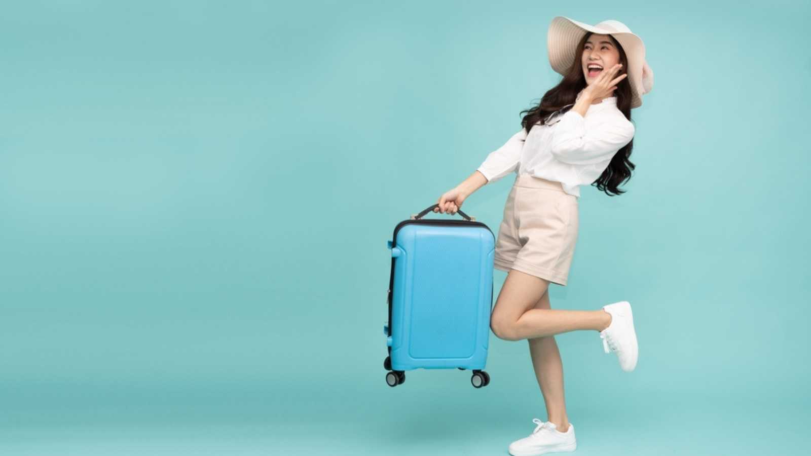 Happy Asian woman traveler standing and holding suitcase isolated on green background, Tourist girl having cheerful holiday trip concept, Full body composition