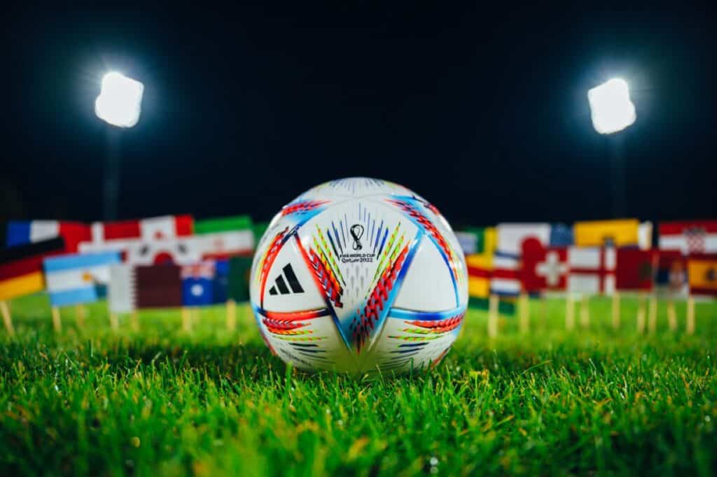 Official Adidas World Cup Football Ball Al Rihla on green grass. Flags of all 32 nations participate on FIFA World Cup in Qatar 2022. Soccer Ball at Stadium in night