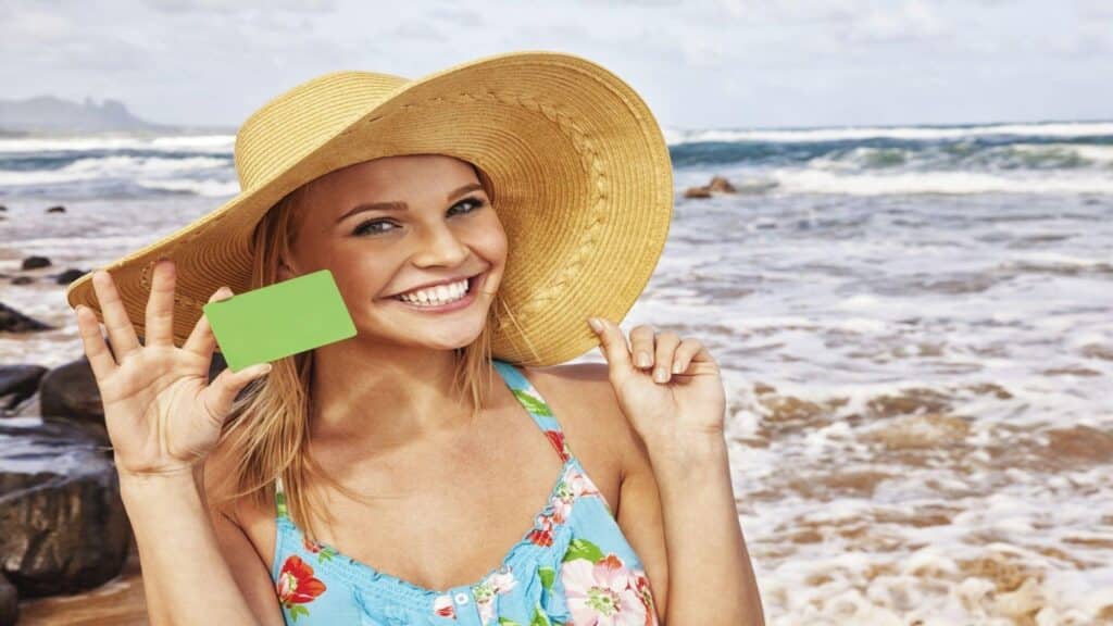 Photo of an attractive, happy young blonde woman on a Hawaiian beach, wearing a large floppy beach hat, holding a blank green credit card (or gift, reward, incentive, etc.).