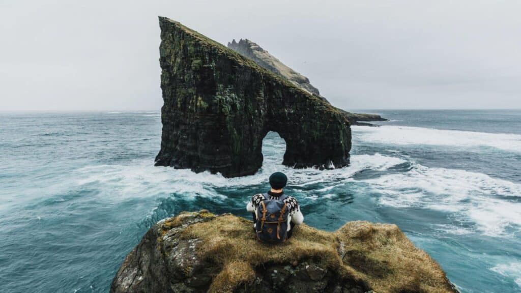 Young Caucasian man in knitted sweater looking at view of Drangarnir arch in Faroe Islands