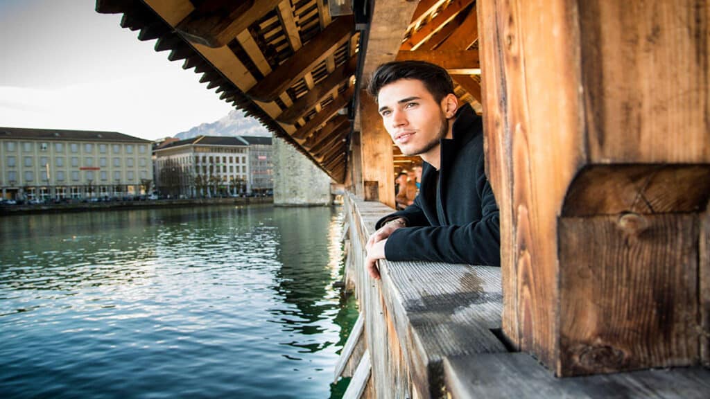 Young dark haired man standing on wooden medieval footbridge in Switzerland while looking away