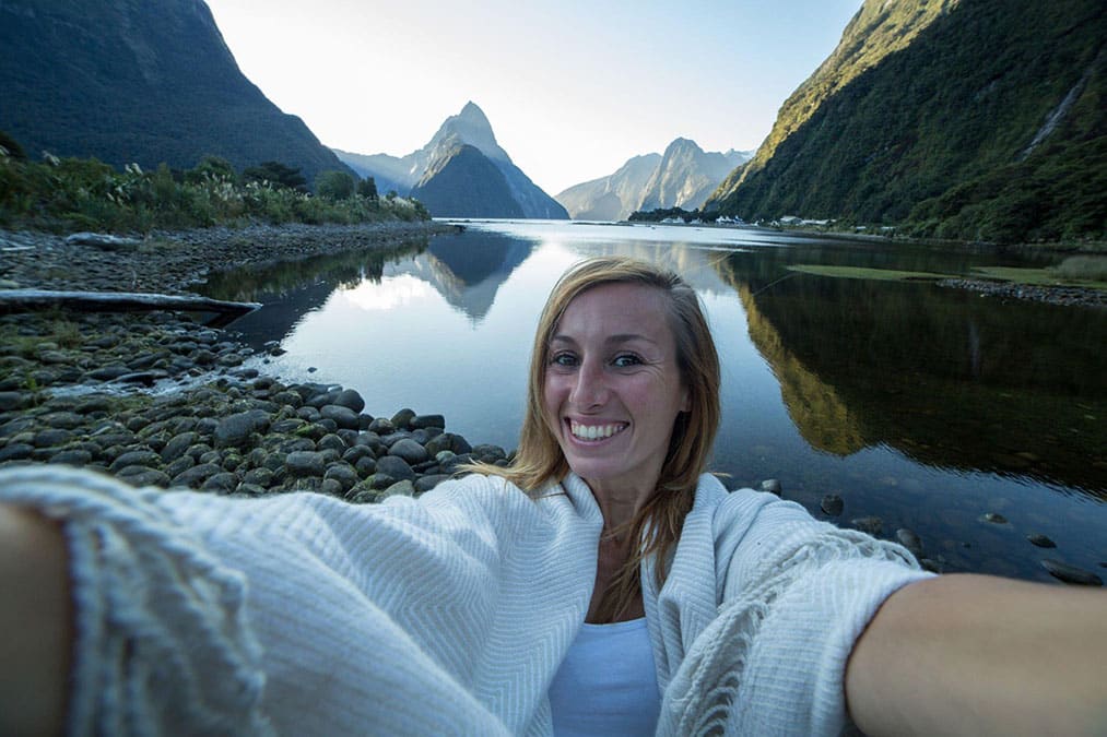 Cheering young woman takes a selfie portrait in front of the Mitre peak in Milford sound, New Zealand.