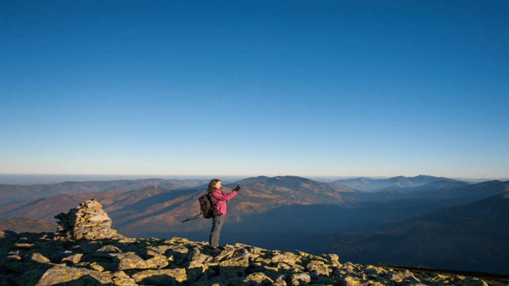 Young beautiful girl standing on rocky mountain top and taking picture with her smartphone.